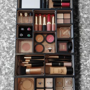 May include: A black and gold makeup and jewellery organiser, filled with cosmetics and jewellery. The organiser contains lipsticks, eyeshadow palettes, blush, makeup brushes, foundation, perfumes, rings, and necklaces. The colour scheme includes black, gold, and various makeup shades.
