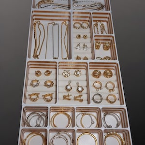Puede incluir: Un organizador de joyas lleno de varios collares, pulseras, anillos y pendientes de oro y plata. Las joyas están dispuestas en una bandeja blanca con divisores a rayas doradas, mostrando una variedad de estilos y diseños.