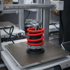 Può includere: Un portapenne a forma di molla rossa e nera con due penne all'interno, appoggiato su una stampante 3D. Il portapenne ha un design a spirale e una base rotonda. La stampante è bianca e grigia.