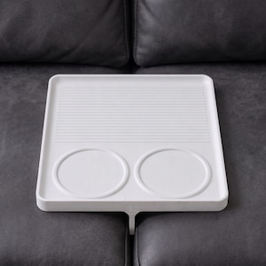 Puede incluir: Una bandeja de sofá blanca y rectangular con una superficie superior acanalada y dos huecos circulares para tazas. La bandeja tiene una sola pata central que se encaja entre los cojines del sofá. Ideal para bebidas y aperitivos.