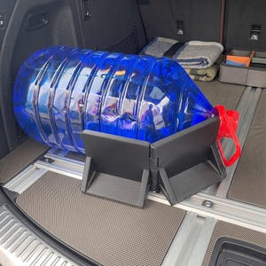 May include: A large, transparent blue water jug with a red handle and cap, resting on a black support structure inside a vehicle's cargo area. The support is designed to hold the jug securely. The cargo area has a gray floor.