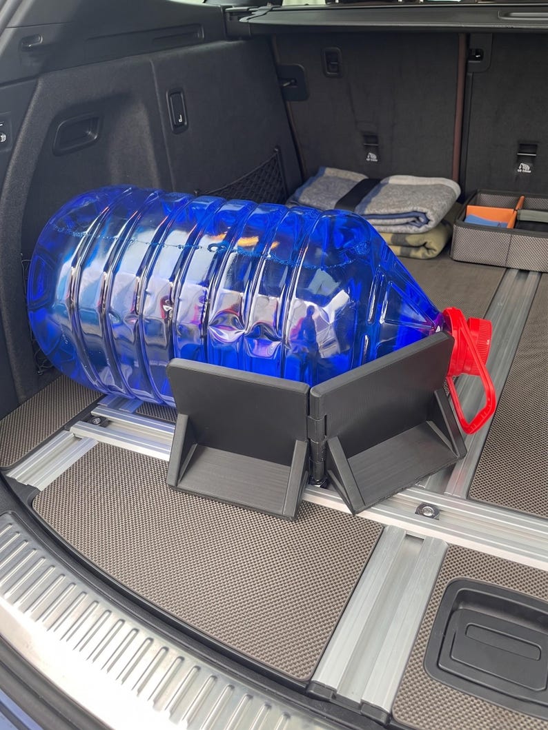 May include: A large, transparent blue water jug with a red handle and cap, resting on a black support structure inside a vehicle's cargo area. The support is designed to hold the jug securely. The cargo area has a gray floor.
