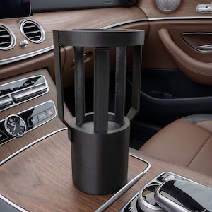 Könnte beinhalten: Ein schwarzer Getränkehalter für das Auto mit zylindrischer Form und geschlitztem Design. Er befindet sich in der Mittelkonsole eines Autos, mit braunen Ledersitzen und Holzzierleisten im Hintergrund.