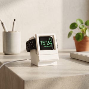 May include: A white watch charging stand with a black watch displaying the time 15:24. The watch has a black strap. A white cable is connected to the stand. A small potted plant and a pen holder are in the background.
