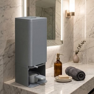 May include: A grey laundry detergent dispenser with a pull-out drawer and a measuring cup. The dispenser is on a marble countertop, next to a rolled grey towel, soap, and a brown soap dispenser.