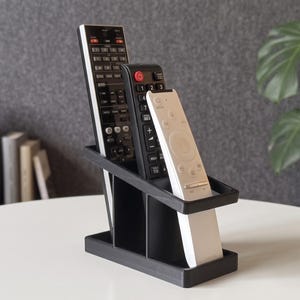 Può includere: Un organizer per telecomandi nero che contiene tre telecomandi. L'organizer è realizzato in materiale nero opaco e ha tre scomparti. I telecomandi sono neri e bianchi, con vari pulsanti e design. L'organizer è su un tavolo bianco.