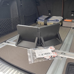 May include: A black, foldable cargo organizer is the focal point, positioned in the trunk of a vehicle. The organizer is accompanied by a small bag of mounting tape. A folded blanket and other items are visible in the background.