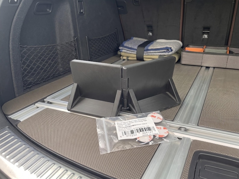 May include: A black, foldable cargo organizer is the focal point, positioned in the trunk of a vehicle. The organizer is accompanied by a small bag of mounting tape. A folded blanket and other items are visible in the background.
