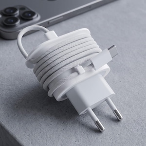 Può includere: Un set di cavo e adattatore di ricarica bianchi, ordinatamente arrotolato e fissato. Il cavo è avvolto attorno all'adattatore, con un connettore USB-C visibile. Uno smartphone sullo sfondo, che suggerisce un accessorio tecnologico.