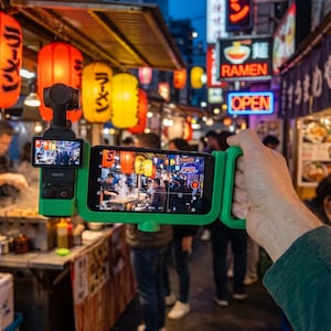 May include: A green smartphone holder with a built-in camera is capturing a bustling street scene at night. The scene includes illuminated ramen shop signs and colorful paper lanterns. The word "OPEN" is visible in neon.