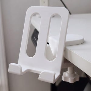 Puede incluir: Soporte para teléfono blanco, impreso en 3D, con una abrazadera para fijar a un escritorio. El soporte tiene un marco rectangular con dos aberturas y una base para sostener el teléfono. La abrazadera tiene un tornillo para ajustar.