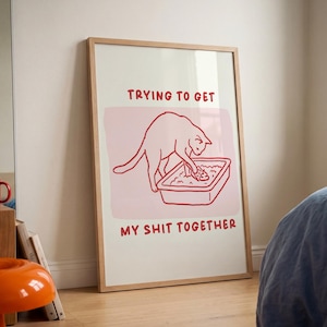 May include: Framed art print featuring a red line drawing of a cat in a litter box, with the text "TRYING TO GET MY SHIT TOGETHER" in red. The artwork is set against a pink background.