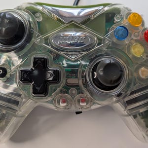 May include: Clear plastic video game controller with black joysticks, a directional pad, and colored buttons. The controller has the brand name "idtec" in silver lettering. The design allows a view of the internal components.