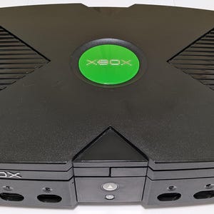 May include: Black Xbox video game console with a green circular power button. The console has a ridged top and the word "XBOX" is printed on the front. A classic gaming console.
