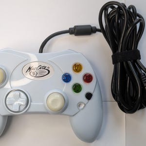 May include: A light blue Mad Catz video game controller with a long black wired cable. The controller features a central logo, a directional pad, two analog sticks, and four colored action buttons labeled X, Y, A, and B.