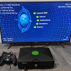 May include: A black original Xbox game console with a matching wired controller, displayed in front of a television screen showing the UnleashX dashboard interface with system information, including CPU and motherboard temperatures in Fahrenheit.