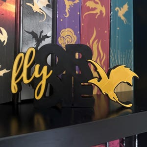 Pode incluir: Uma placa impressa em 3D nas cores preta e amarela com a palavra fly em letra cursiva e as palavras OR DIE em letras de forma, acompanhada por uma silhueta de dragão amarela. A placa está posicionada à frente de livros com lombadas de dragão.