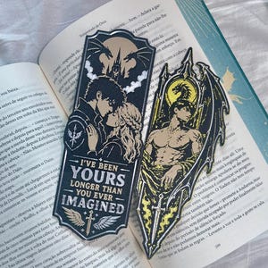 Pode incluir: Dois marcadores de livros decorativos com arte de fantasia. Um apresenta um casal com o texto "I'VE BEEN YOURS LONGER THAN YOU EVER IMAGINED". O outro mostra uma figura alada com uma espada, em amarelo e preto.