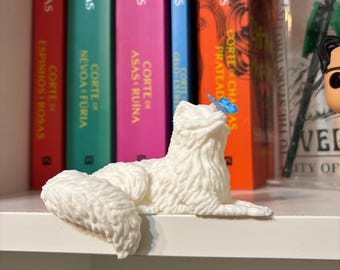 Può includere: Una statuetta di gatto bianco e soffice con una farfalla blu sul naso. Il gatto è sdraiato su una mensola bianca, con libri colorati sullo sfondo. I libri hanno titoli in spagnolo e portoghese.