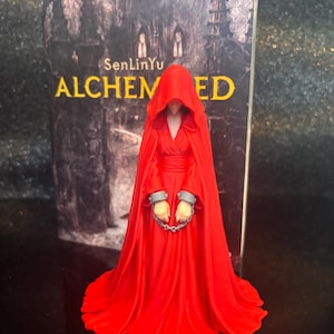 Puede incluir: Una figura con una larga y fluida capa roja con capucha y vestido, con grilletes grises en las mu&ntilde;ecas unidos por una cadena corta. La figura se encuentra frente a la portada de un libro titulada SenLinYu ALCHEMIZED con una imagen de un bosque.