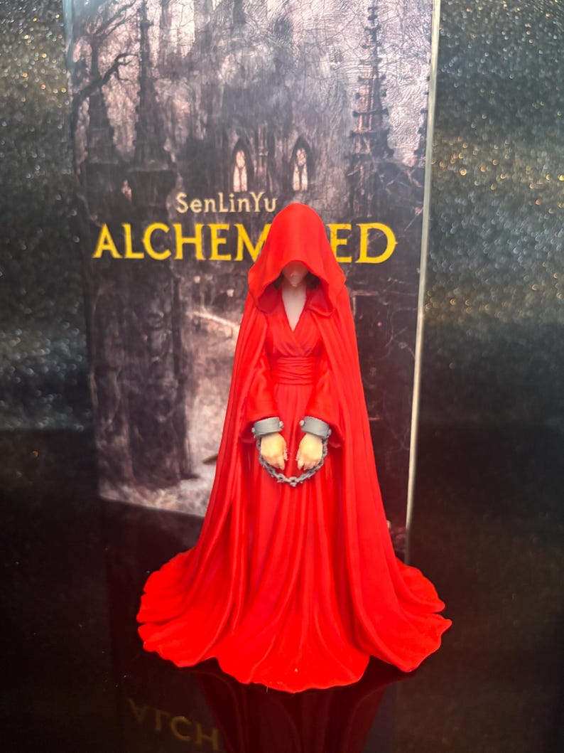 Puede incluir: Una figura con una larga y fluida capa roja con capucha y vestido, con grilletes grises en las mu&ntilde;ecas unidos por una cadena corta. La figura se encuentra frente a la portada de un libro titulada SenLinYu ALCHEMIZED con una imagen de un bosque.