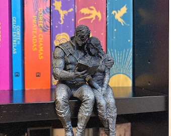May include: A gray, textured figurine of two figures sitting together, with one figure holding an open book while the other leans against them. The sculpture features detailed clothing and hair, capturing a moment of shared reading in a seated pose.