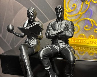 Può includere: Due statuette nere stampate in 3D sedute su una sporgenza. Una figura tiene in mano una sfera e un libro aperto, mentre l'altra è seduta con le braccia incrociate. Dietro c'è una cornice rettangolare gialla con una sagoma di drago nero.