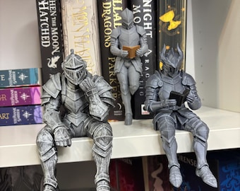 Può includere: Tre statuette di cavalieri grigi in armatura completa sono esposte su uno scaffale. Un cavaliere è seduto a sinistra con una mano sul mento, un altro è in piedi al centro che legge un libro e il terzo è seduto a destra che legge un libro.
