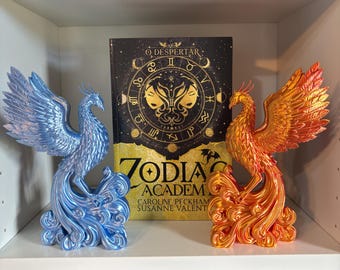 Può includere: Due statuette di fenice, una blu metallizzato e una arancione metallizzato, si trovano su uno scaffale ai lati di un libro intitolato Zodiac Academy: O Despertar. Gli uccelli hanno piume dettagliate e code a volute. La copertina mostra uno zodiaco.
