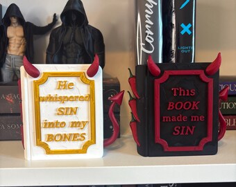 Può includere: Due oggetti rettangolari a forma di libro con corna e code da diavolo. Quello bianco presenta la scritta dorata "He whispered SIN into my BONES". Quello nero presenta la scritta rossa "This BOOK made me SIN" e decorazioni a peperoncino.