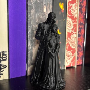 Puede incluir: Una figura negra mate impresa en 3D que representa a dos personas de pie en un abrazo. La figura m&aacute;s alta lleva un abrigo largo, mientras que la m&aacute;s baja tiene el pelo largo y rizado y un vestido hasta el suelo. La escultura est&aacute; erguida.