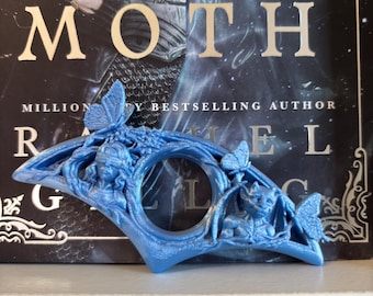 Può includere: Un ferma pagine blu con una figura scolpita con una benda sugli occhi a sinistra e una creatura alata a destra, decorato con farfalle. L'oggetto è posizionato davanti a una copertina di libro con il testo visibile KNIGHT AND THE MOTH.