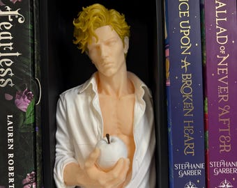 Può includere: Un busto maschile con capelli gialli ricci e carnagione pallida, che indossa una camicia bianca sbottonata. La figura tiene in mano una piccola mela bianca ed è esposta all'interno di una cornice rettangolare nera tra due libri.
