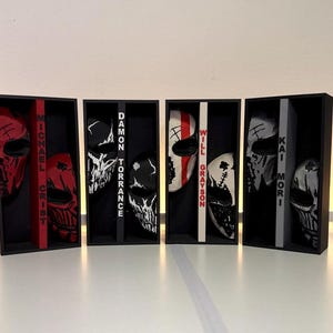 May include: Four rectangular black display boxes, each containing two stylized masks and a central vertical spine. The spines feature the names Michael Crist, Damon Torrance, Will Grayson, and Kai Mori in bold lettering. The masks display intricate graphic patterns in red, black, and white, with each set color-coordinated to match the specific name printed on the spine of its respective display box.