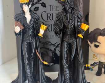 Può includere: Due statuette stampate in 3D di un personaggio maschile con orecchie a punta, che indossa un cappotto nero, pantaloni e una corona dorata. Ogni figura tiene in mano un piccolo libro e un calice dorato. Una figura ha la pelle chiara, l'altra è nera. Sullo sfondo c'è il libro The Cruel Prince.