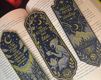 Può includere: Tre segnalibri blu scuro con illustrazioni in lamina d'oro e argento. Uno riporta la scritta "TO THE STARS WHO LISTEN, AND THE DREAMS THAT ARE ANSWERED.", un altro "THE SURIEL HAS tea.", e il terzo "HELLO, FEYRE darling." con motivi di montagne.