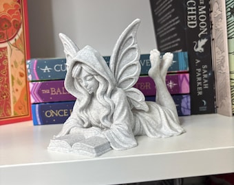 Può includere: Una statuetta grigio chiaro testurizzata raffigurante una fata con ali, che indossa una veste con cappuccio, sdraiata sulla pancia mentre legge un libro aperto. La fata ha i capelli lunghi e i piedi sollevati dietro.