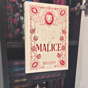 Peut inclure: Un livre relié intitulé God of Malice de Rina Kent avec une couverture crème ornée de bordures rouges, de roses, d'une icône de masque central avec des yeux en X, et de petites illustrations d'un cœur, d'un crâne, d'un appareil photo et de ciseaux.