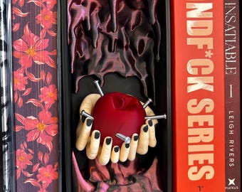 Può includere: Un inserto per libreria stampato in 3D raffigurante un paio di mani pallide con unghie dipinte di nero che reggono un cuore rosso trafitto da quattro chiodi argentati, su uno sfondo scuro, lucido e gocciolante in una cornice.