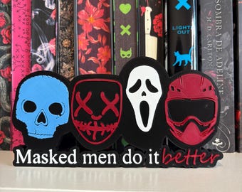 Può includere: Un cartello nero stampato in 3D con quattro maschere distinte: un teschio blu, una maschera rossa con cuciture, una maschera da fantasma bianca e un casco rosso. Il testo "Masked men do it better" è stampato in bianco e rosso alla base.