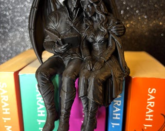 Può includere: Una statuetta nero opaco di due figure alate sedute insieme, con una figura che legge un libro mentre l'altra si appoggia a essa. Le figure indossano abiti dettagliati e sono appollaiate sopra una fila di libri di Sarah J. Maas.
