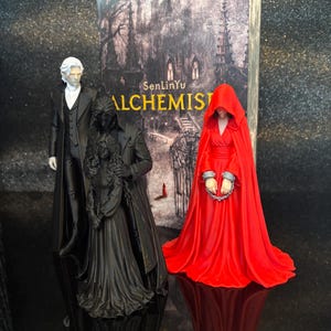 Puede incluir: Tres figuras se encuentran frente a un libro titulado SenLinYu ALCHEMIST. Una figura viste traje negro y camisa blanca. Otra es una figura vestida de negro que sostiene a una persona con vestido largo. La tercera lleva una capa roja con capucha y grilletes.