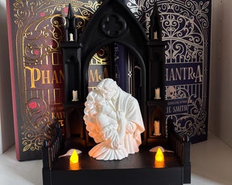 Può includere: Una struttura ad arco gotico nero con una scultura bianca di una coppia che si abbraccia. La base include due luci arancioni brillanti e piccole figure di topi bianchi. L'oggetto è posizionato davanti a due libri intitolati Phantom e Mantra.