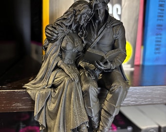 Puede incluir: Una figura impresa en 3D de color negro mate que muestra a dos personajes sentados juntos. Un personaje con cabello largo y orejas puntiagudas apoya la cabeza en el hombro del otro, que sostiene un libro abierto. Ambos llevan capas y botas detalladas.