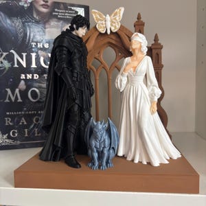 Puede incluir: Una figura impresa en 3D que muestra a un personaje con armadura negra y otro con los ojos vendados y vestido blanco frente a un arco gótico marrón con una mariposa. Una pequeña gárgola gris se sitúa entre ambos sobre una base rectangular.