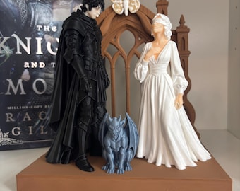 Può includere: Una statuetta stampata in 3D con una figura in armatura nera e una figura bendata in abito bianco davanti a un arco gotico marrone con una farfalla. Un piccolo gargoyle grigio è posizionato tra loro su una base rettangolare marrone.