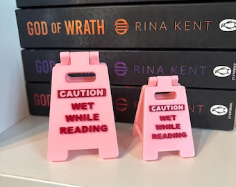 Può includere: Due cartelli di avvertimento in plastica rosa chiaro di dimensioni diverse, ciascuno con la scritta CAUTION WET WHILE READING in lettere rosso scuro. I cartelli hanno la forma di cavalletti da pavimento in miniatura e sono davanti ai libri.