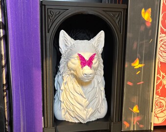 Puede incluir: Un busto de lobo blanco colocado dentro de un marco negro arqueado. Una mariposa de color rosa brillante está adherida al puente de la nariz del lobo. El lobo tiene una textura de pelaje detallada y orejas puntiagudas, mientras que el marco presenta sutiles detalles de hojas talladas.