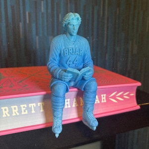Op de afbeelding: Blauw beeldje van een hockeyspeler die een boek leest, zittend op een roze en blauw boek. De speler draagt een trui met "Briar 44". De rug van het boek luidt "Brett Hannah". De achtergrond is een blauwe en zwarte textuur.