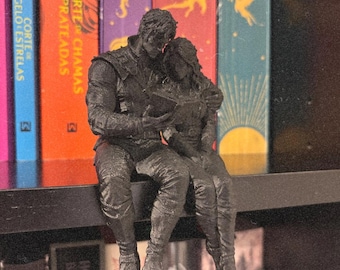 Può includere: Una statuetta stampata in 3D nero opaco che raffigura due figure sedute insieme, con una che abbraccia l'altra mentre entrambe tengono e guardano un libro aperto. La scultura testurizzata è progettata per sedersi sul bordo di uno scaffale.